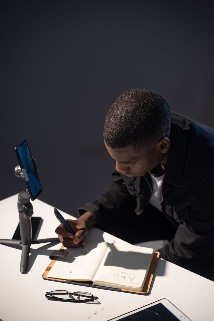 A man concentrates on writing at his desk, using a phone tripod for remote work in Abuja, Nigeria.