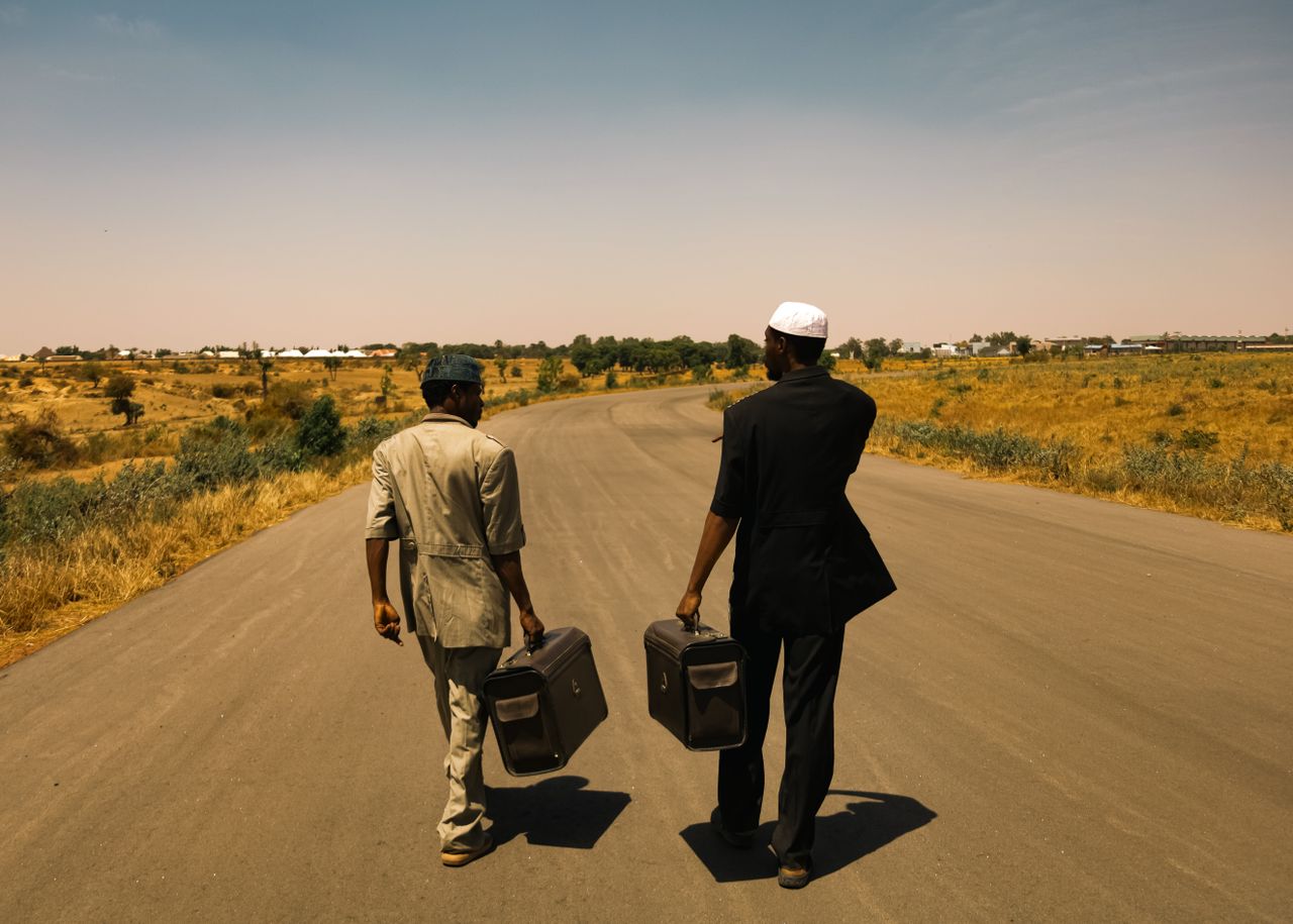 Two men in suits walking down an empty rural road, carrying suitcases under a clear sky.