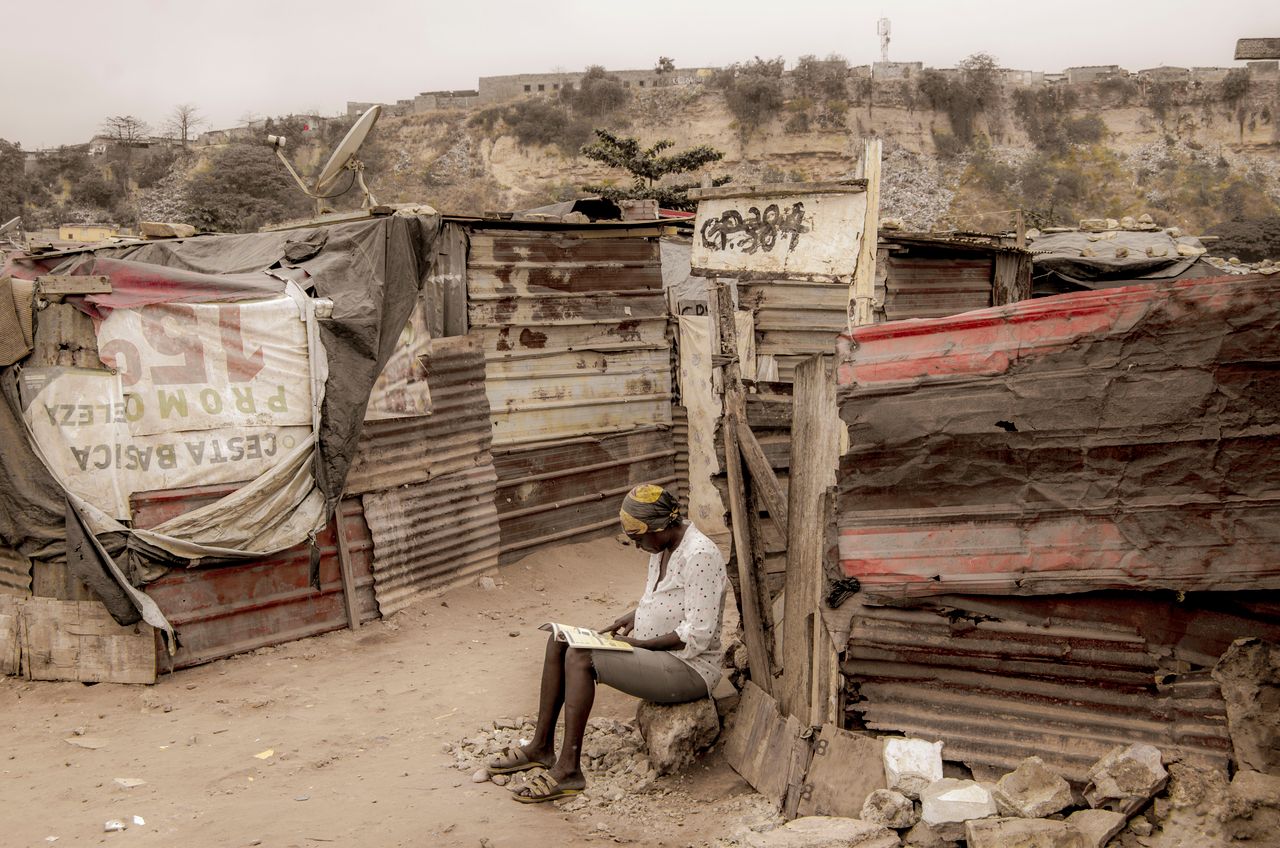 A young girl reads in a makeshift settlement in Luanda, Angola, amidst rustic surroundings.